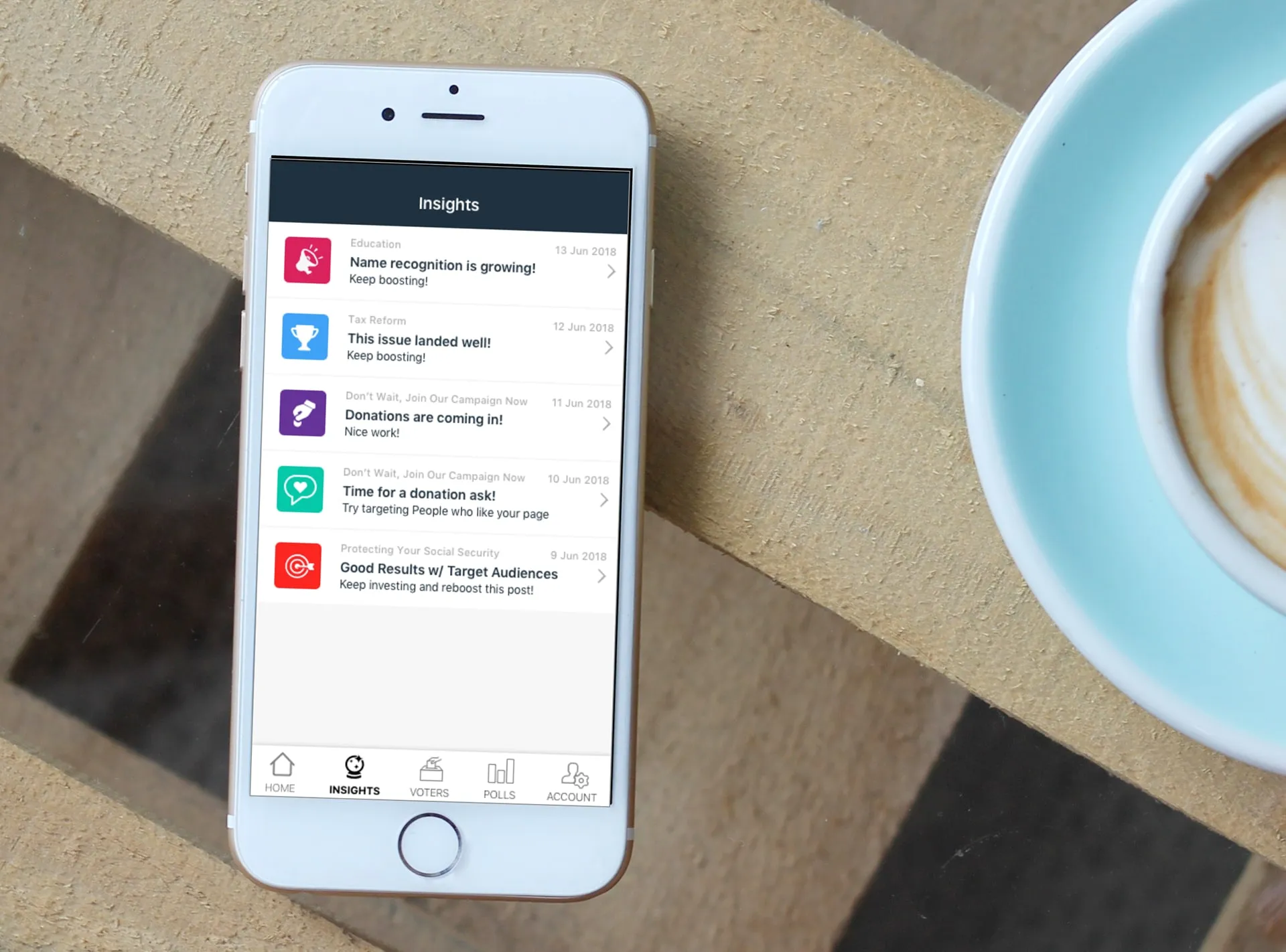 Phone on a wooden table beside a coffee cup, showing the Insights screen with a list of colored icons, short headlines and suggested actions.
