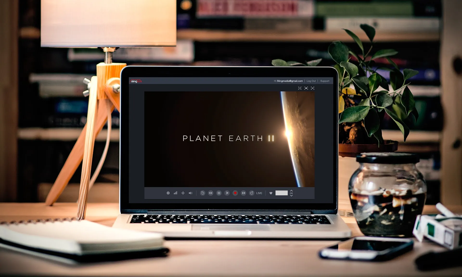 A laptop on a desk with a lamp and plant, showing SlingPlayer playing a Planet Earth II title card with transport controls under the video.