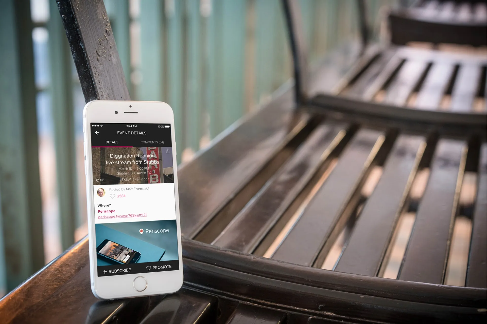 A phone leaning on a dark bench showing an Event Details screen for a live stream event, with a Periscope link and Subscribe and Promote buttons.